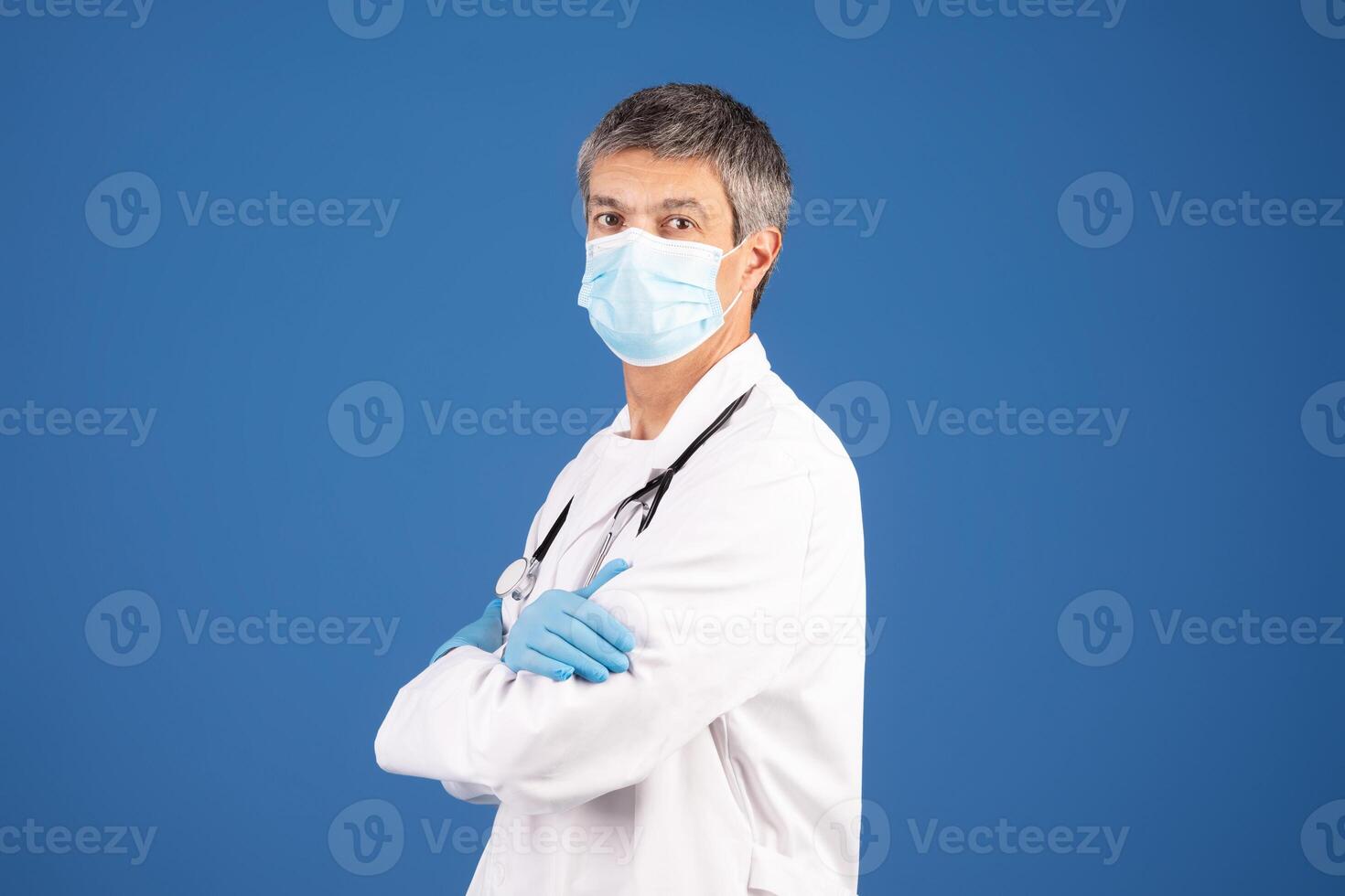 A mature Caucasian man in his 50s, dressed in a white coat, blue gloves, and a mask stands confidently against a solid blue background, representing a committed general physician. photo