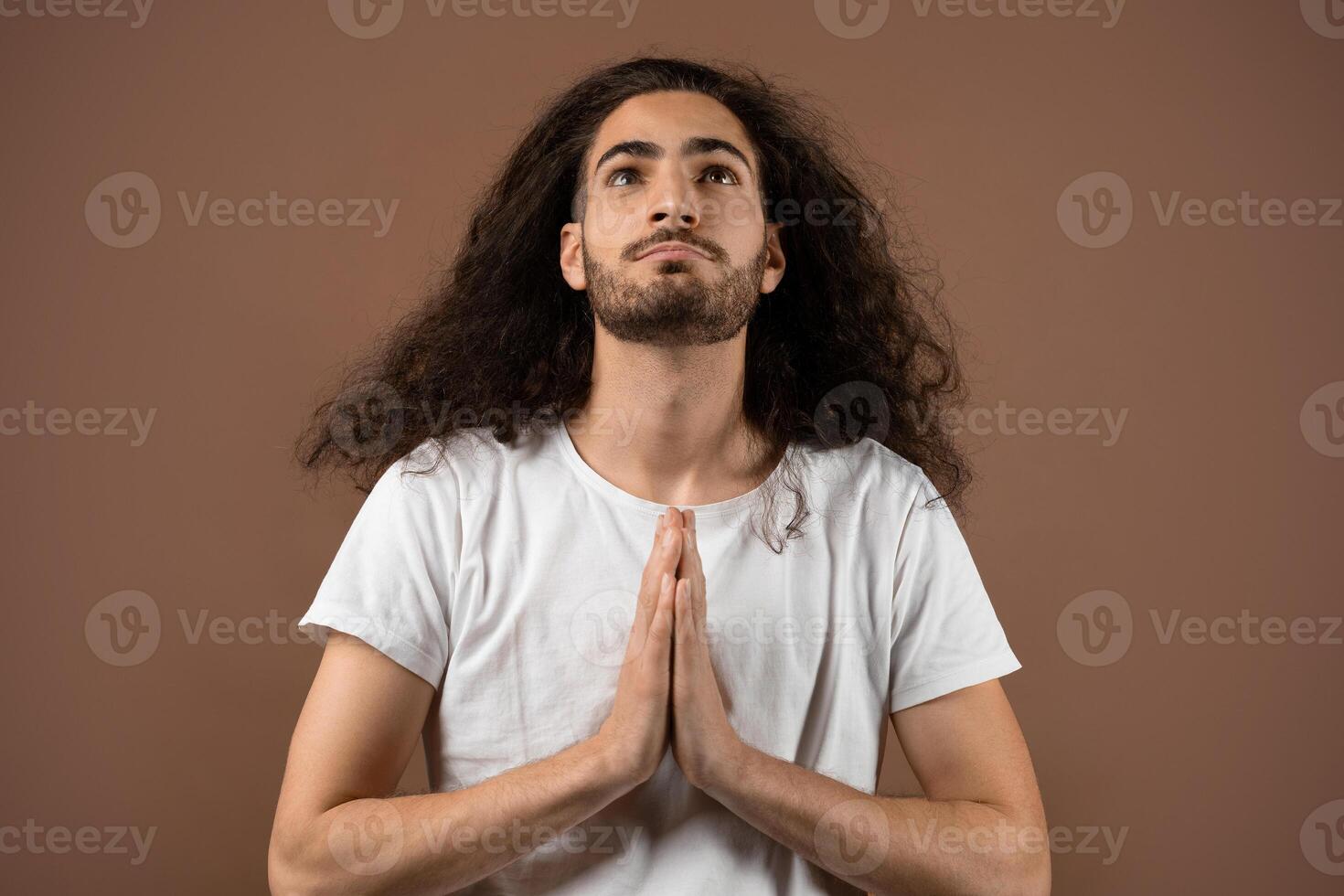 A young Caucasian man with long, curly hair is seen in a tranquil moment, hands together in a gesture of meditation or prayer. His expression reflects calmness and introspection. photo