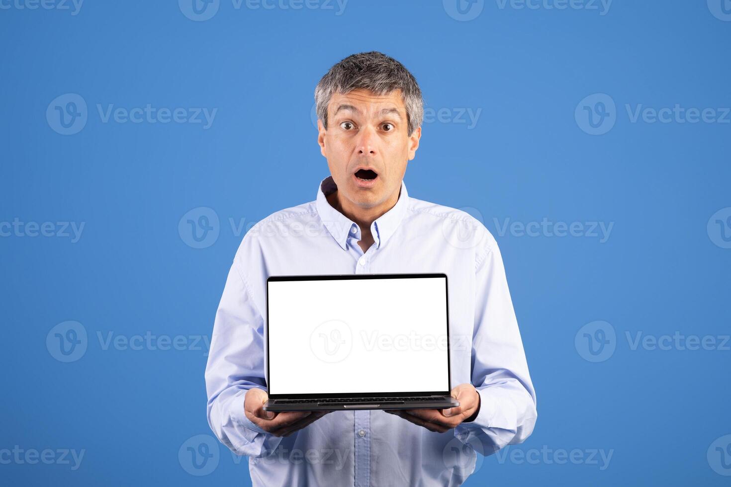 A middle aged Caucasian man stands against a blue background, expressing surprise while holding a blank laptop. His body language reflects astonishment in this isolated moment. photo