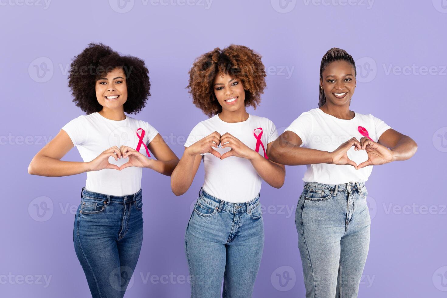 Three young African American women stand together, each forming a heart shape with their hands. They wear matching white shirts and pink ribbons while smiling joyfully against a solid purple backdrop. photo