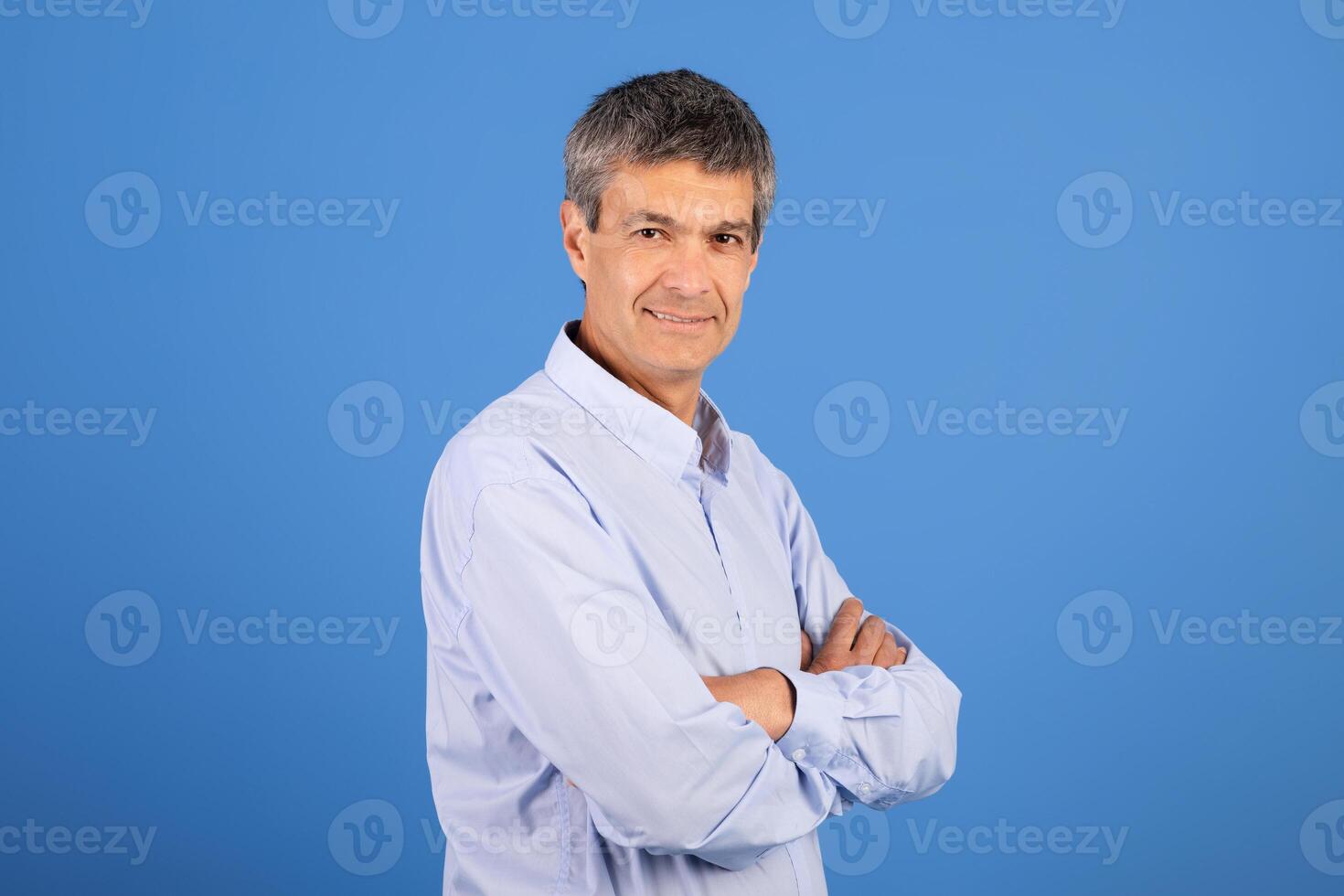 A mature Caucasian man in his 50s stands with arms crossed, smiling gently. He is dressed in a light blue shirt and is surrounded by a simple blue background, creating a relaxed atmosphere. photo