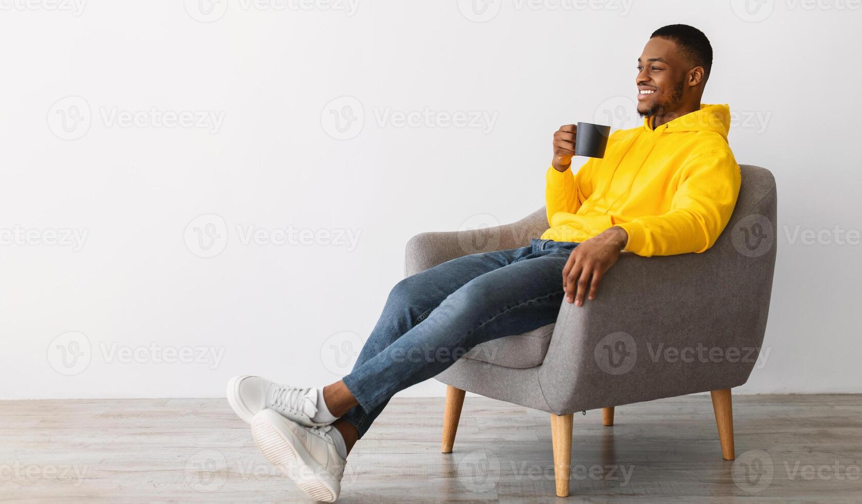 Side View Of Cheerful African American Guy Drinking Coffee Sitting In Chair Over Gray Background. Black Man Enjoying Weekend Relaxing With Hot Drink Indoors, Looking Aside. Panorama photo