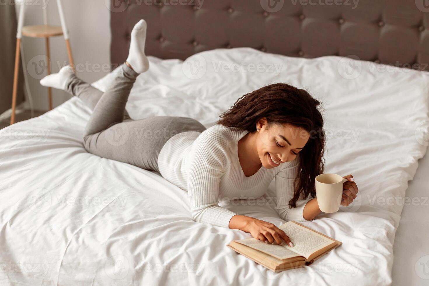 Cozy Weekend. Portrait of smiling African American woman lying in comfortable bed, reading paper book, holding cup of hot coffee, enjoying free time, free copy space, banner. Rest and relax photo