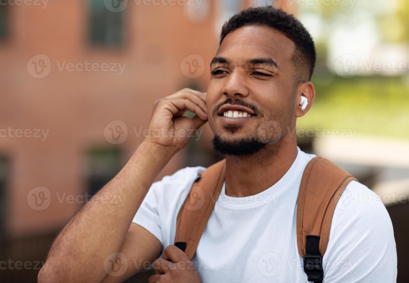 Closeup portrait of handsome black guy wearing earpods and listening to music outdoors, walking in urban city area, free space. Man resting after work and enjoying favorite song photo