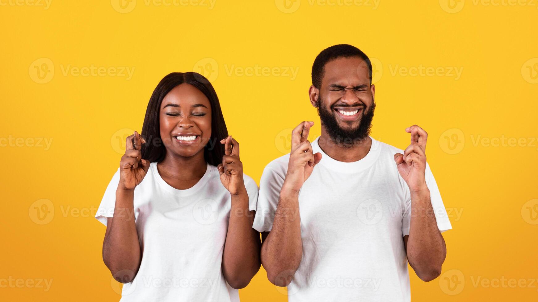 Making Cherished Wish. Positive Black Couple Crossing Fingers And Smiling, Begging For Luck, Praying For Miracle, Hoping For Good Things To Happen, Standing With Closed Eys Over Yellow Studio Wall photo