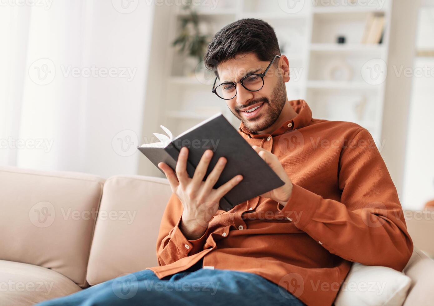 Portrait of young Middle Eastern man wearing eyeglasses holding paper book or diary, sitting on couch at home in living room, reading literature or checking his working schedule plan, free copy space photo