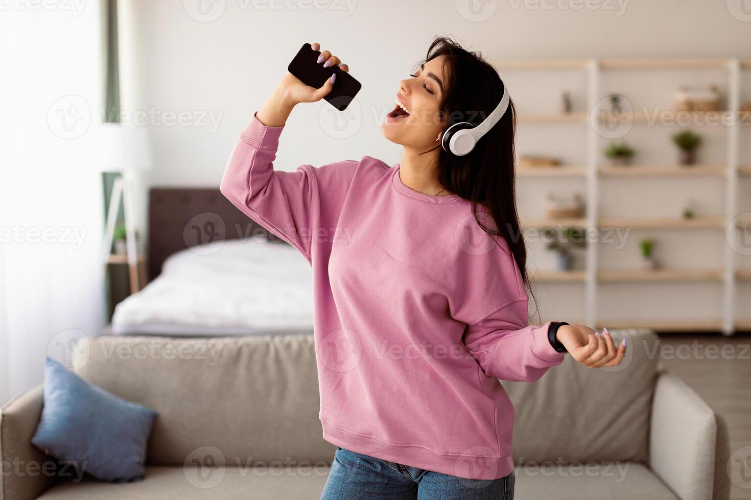 Carefree Singer. Portrait of young lady singing favorite song and using cell phone as a microphone, dancing and wearing wireless headset. Excited woman having fun at home, enjoying sound photo