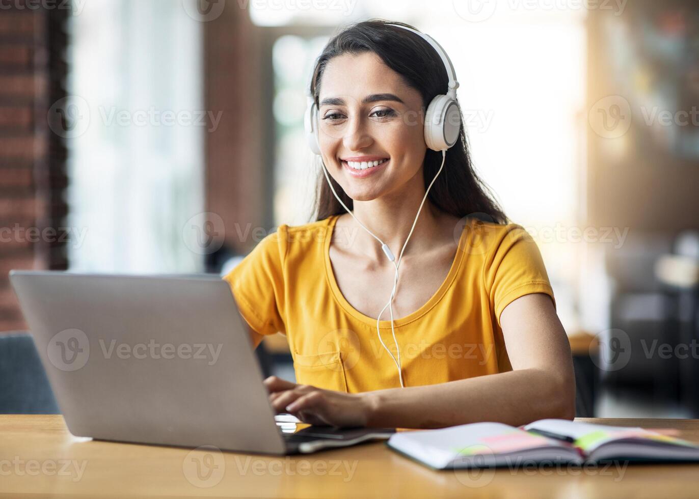 Smiling arab girl with headset studying online, using laptop at cafe, taking notes, copy space photo
