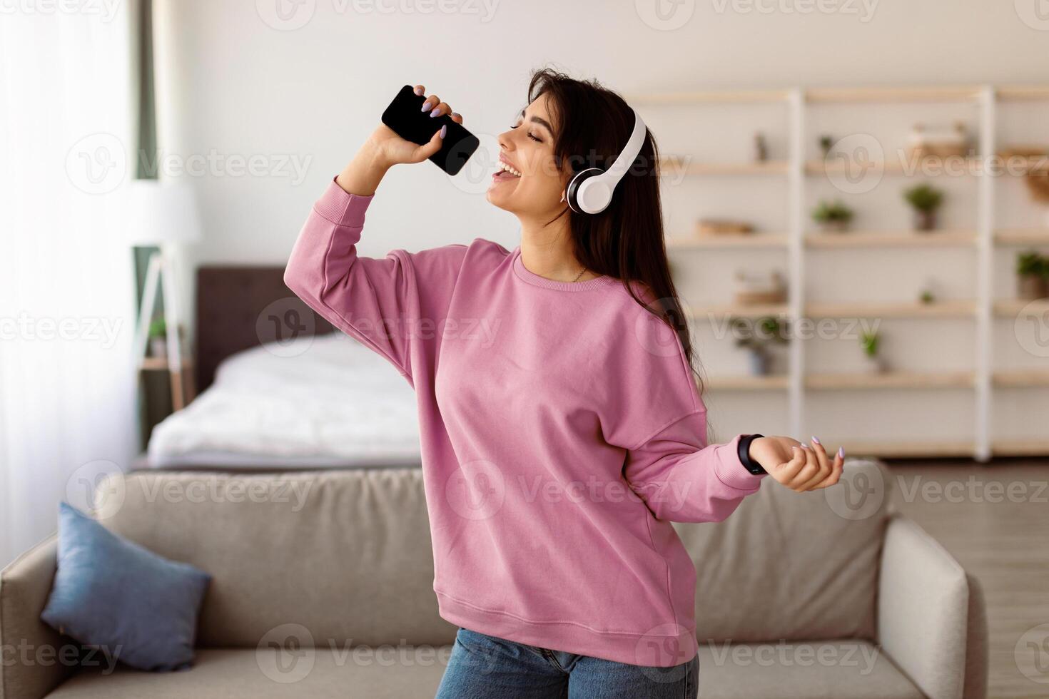 Express Yourself. Cheerful positive young woman singing favorite song in mic, holding and using her smartphone with blank empty screen, dancing in headphones, having fun in living room, relaxing alone photo