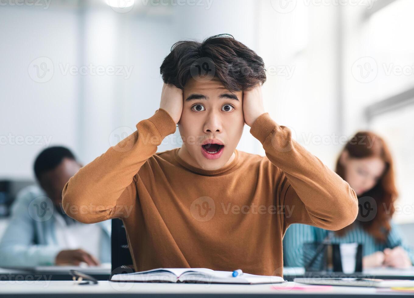 Portrait of shocked asian male student sitting at desk in classroom, grabbing his head looking at camera. Worried youth unprepared for test or exam, thinking about deadline or hard new theme photo