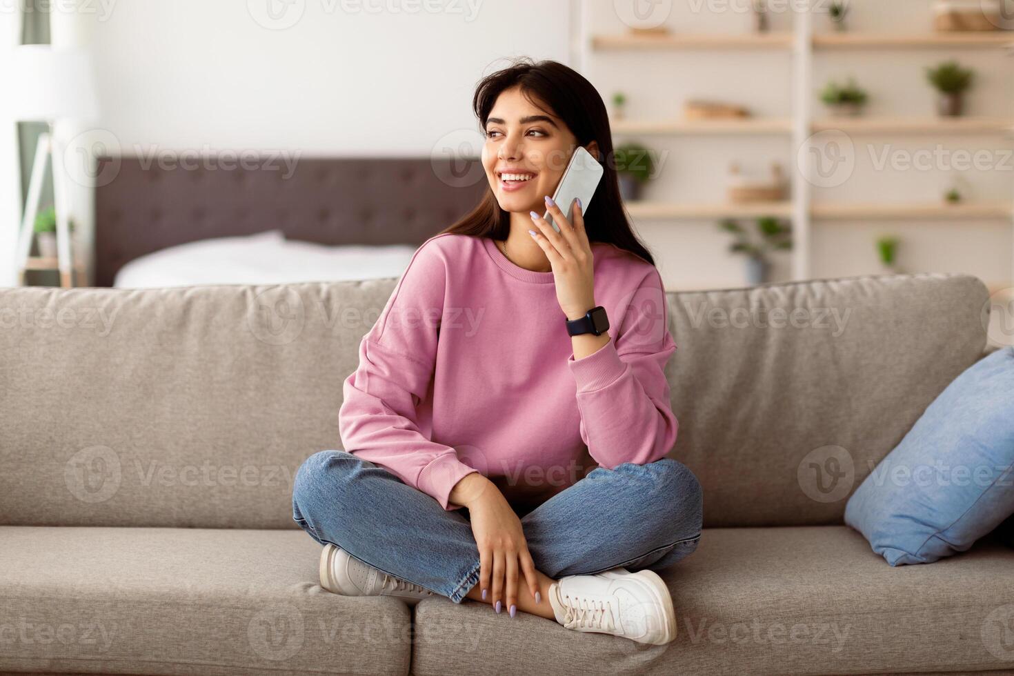 Communication Concept. Portrait of excited smiling young woman talking with friends or family on mobile phone sitting on the couch, looking away, selective focus, blurred background photo