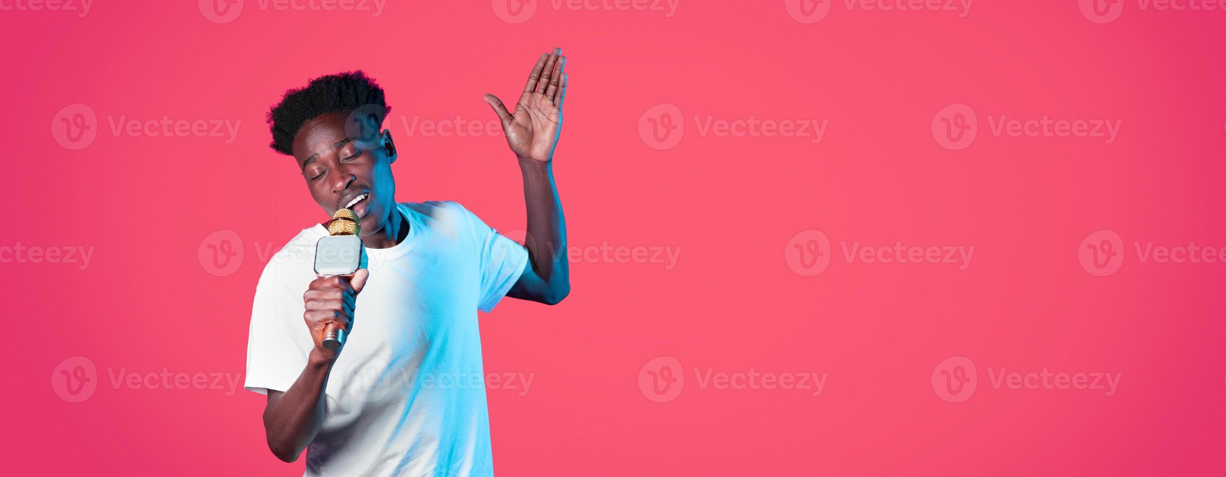 A young black guy entertains with exuberant singing while holding a microphone, showcasing enthusiasm and musical talent. The bright pink backdrop adds to the joyful ambiance. photo
