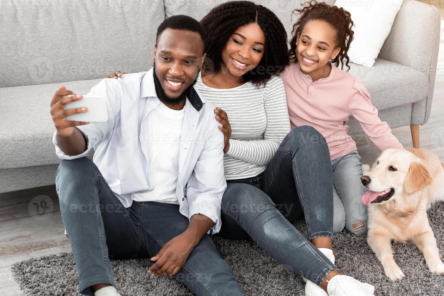Happy Loving Family. Portrait of loving black family taking selfie with their dog in living room, sitting on the floor at home. Man using smartphone for photo, spending time with domestic animal photo
