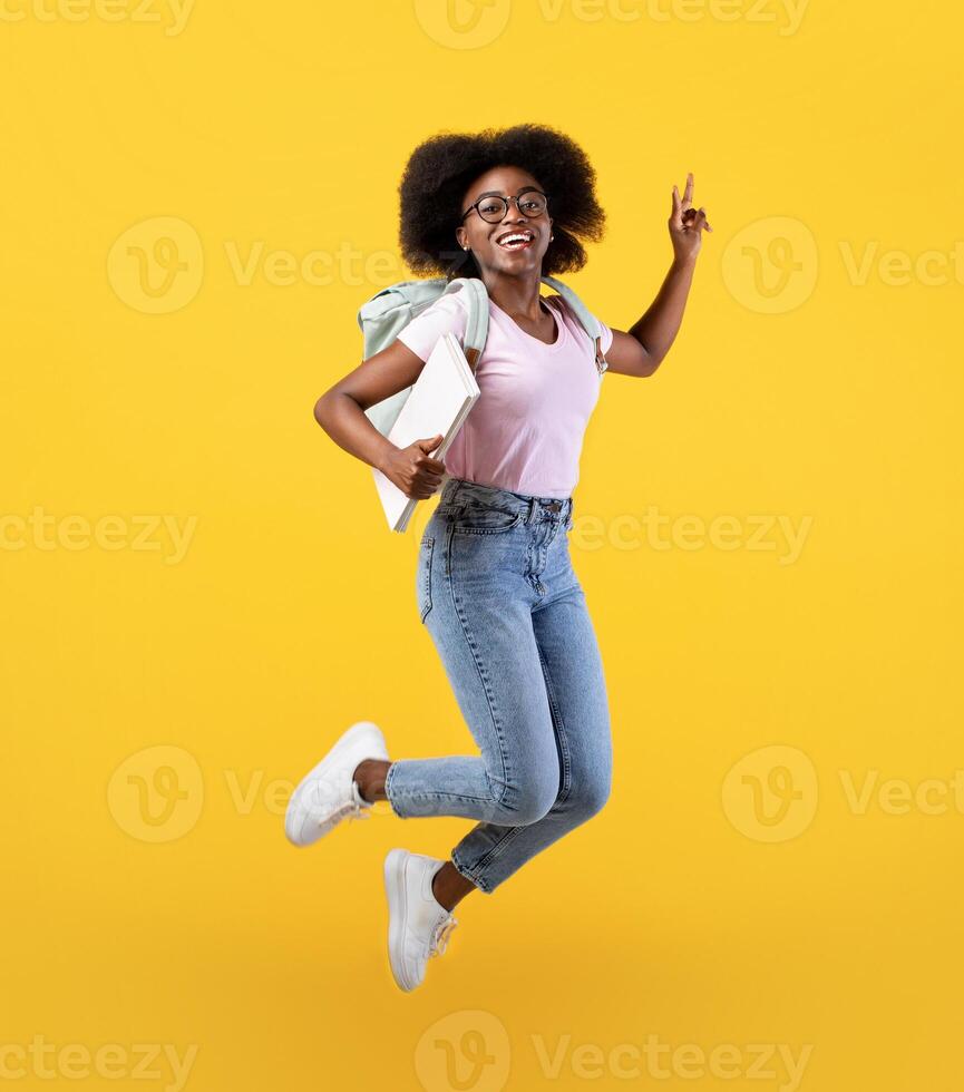 Enjoying student life. Full length portrait of african american lady jumping, wearing backpack and holding notepads over yellow studio background, crop photo