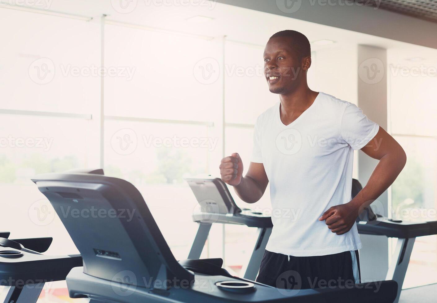 Young happy handsome african-american man in fitness club. Cardio workout, running on treadmill. Healthy lifestyle, guy training in gym. Flare effect, copy space photo