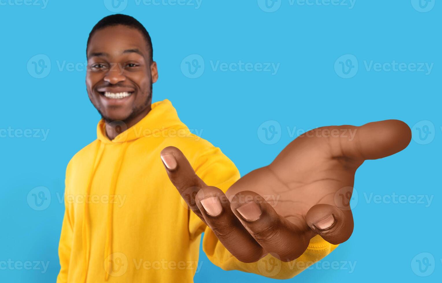 Happy African American guy showing big outstretched hand, offering help, taking or giving something, reaching out for support on blue studio background. Selective focus photo