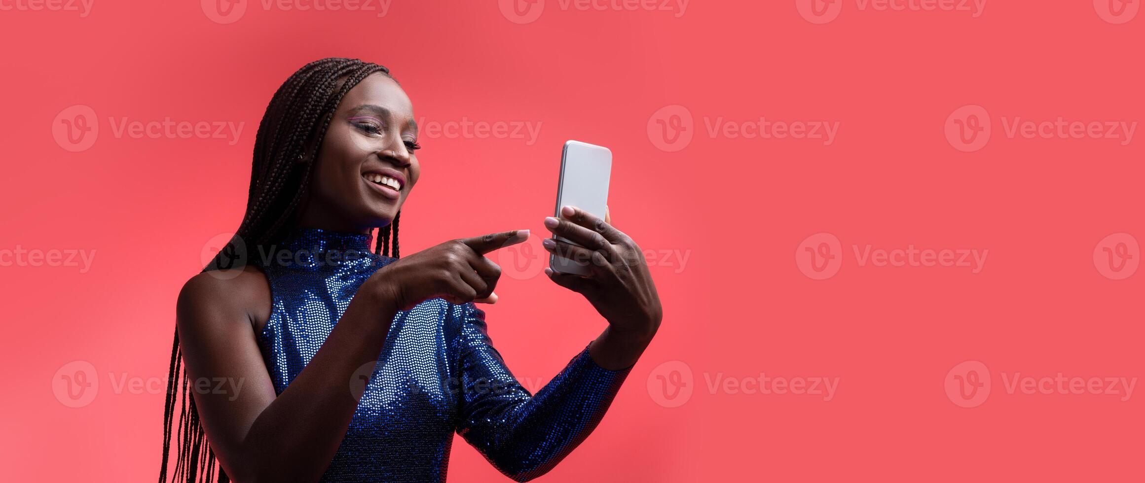 A black woman with braided hair is smiling while looking at her smartphone. She appears engaged and happy, set against a vibrant red background that adds energy to the moment. photo