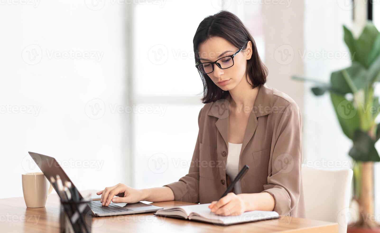 Note Taking. Portrait of female student using laptop and writing memo in her diary at modern office, copyspace photo