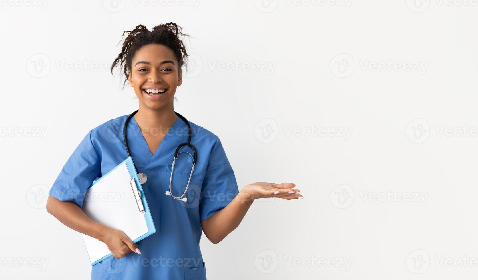 Excited black female doctor in scrubs holding clipboard showing something on empty palm pointing with hand at free copy space, advertising poduct or clinic, posing isolated on white studio background photo