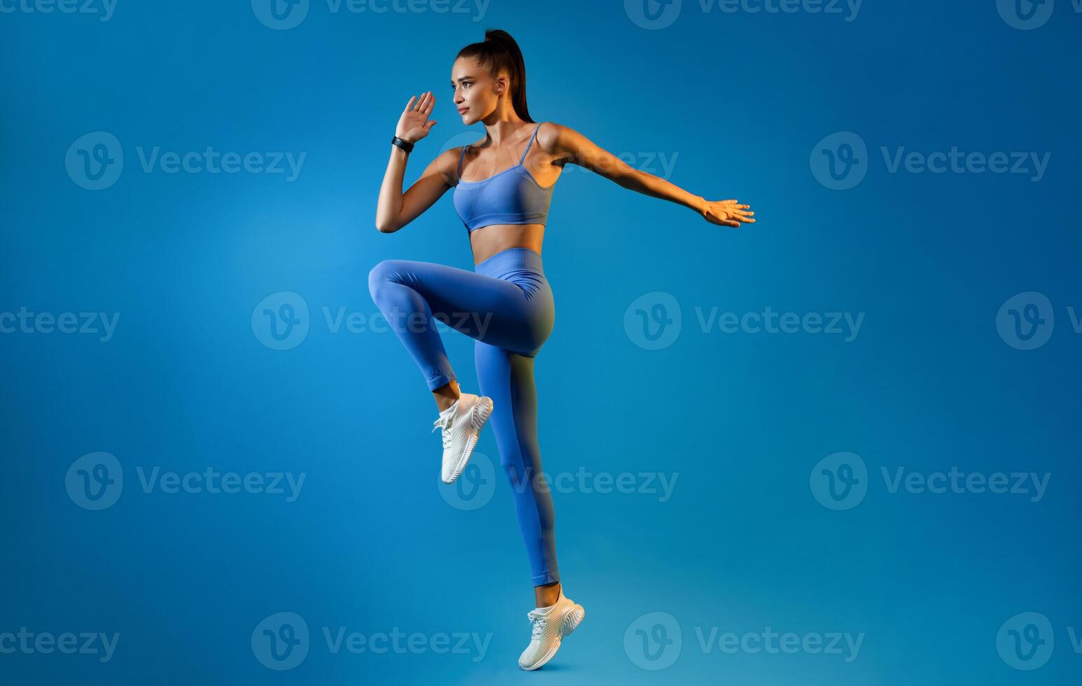 Female Athlete Jumping Exercising During Training Over Blue Studio Background, Looking Aside. Fitness Workout And Sport Motivation Concept. Full Length, Side View photo