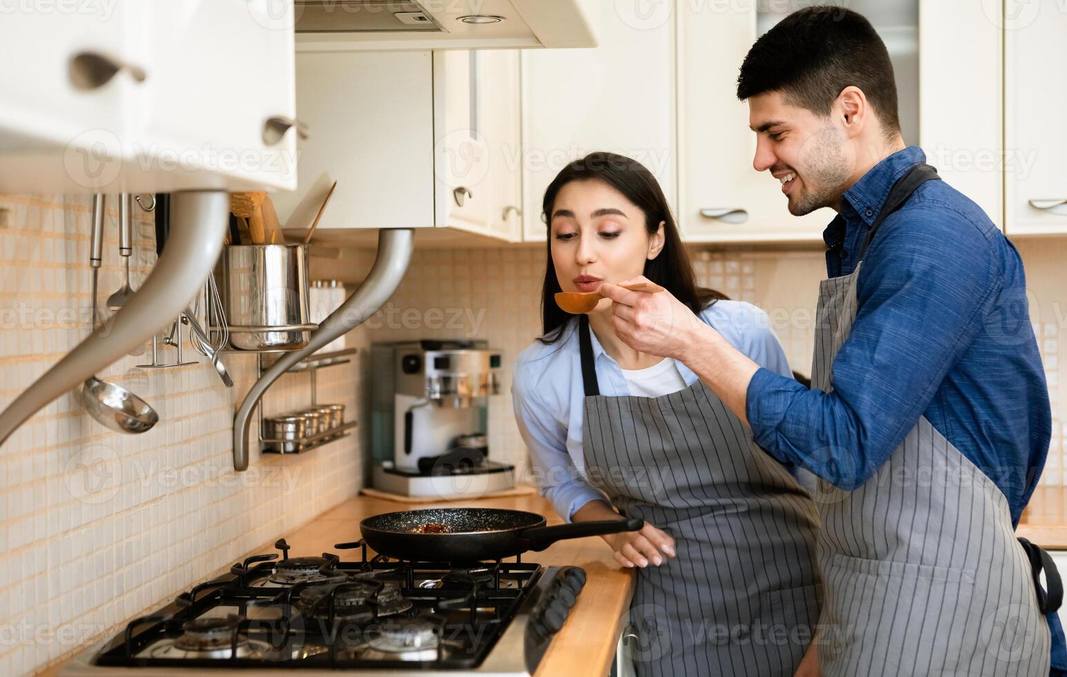 Cooking Together Concept. Smiling man feeding his woman, lady tasting hot fried meal using wooden spoon photo