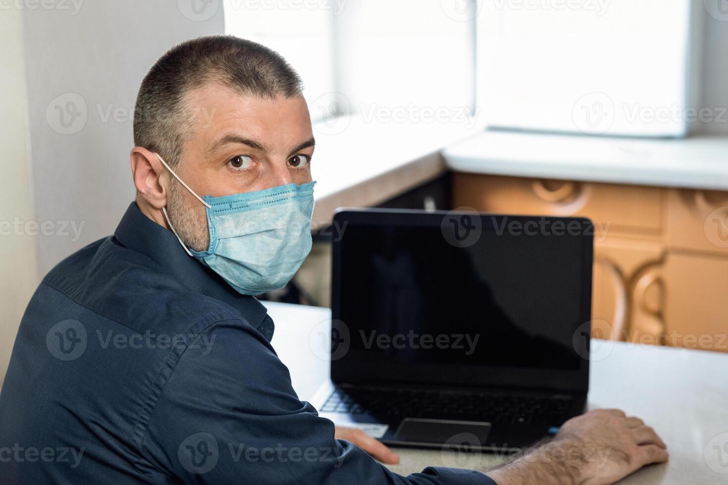 Coronacrisis Concept. Man Sitting At Laptop With Word COVID-19 On Screen, Wearing Medical Mask Working At Home. Selective Focus photo