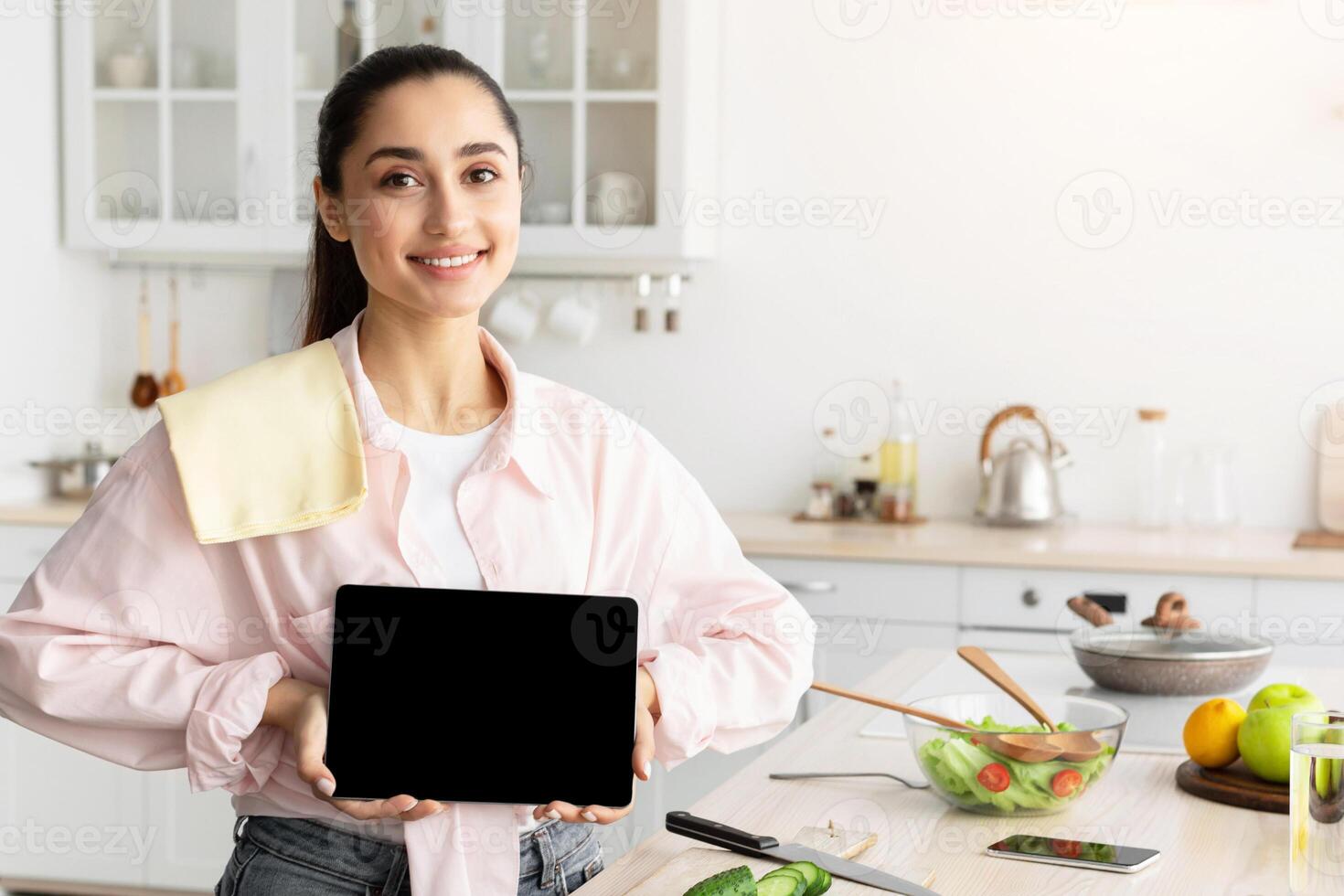 Cheerful Young Lady Showing Empty Digital Tablet Screen Posing Standing In Modern Kitchen At Home. Smiling Woman Recommending Cooking Application Or Website With Healthy Online Recipes. Mock up photo