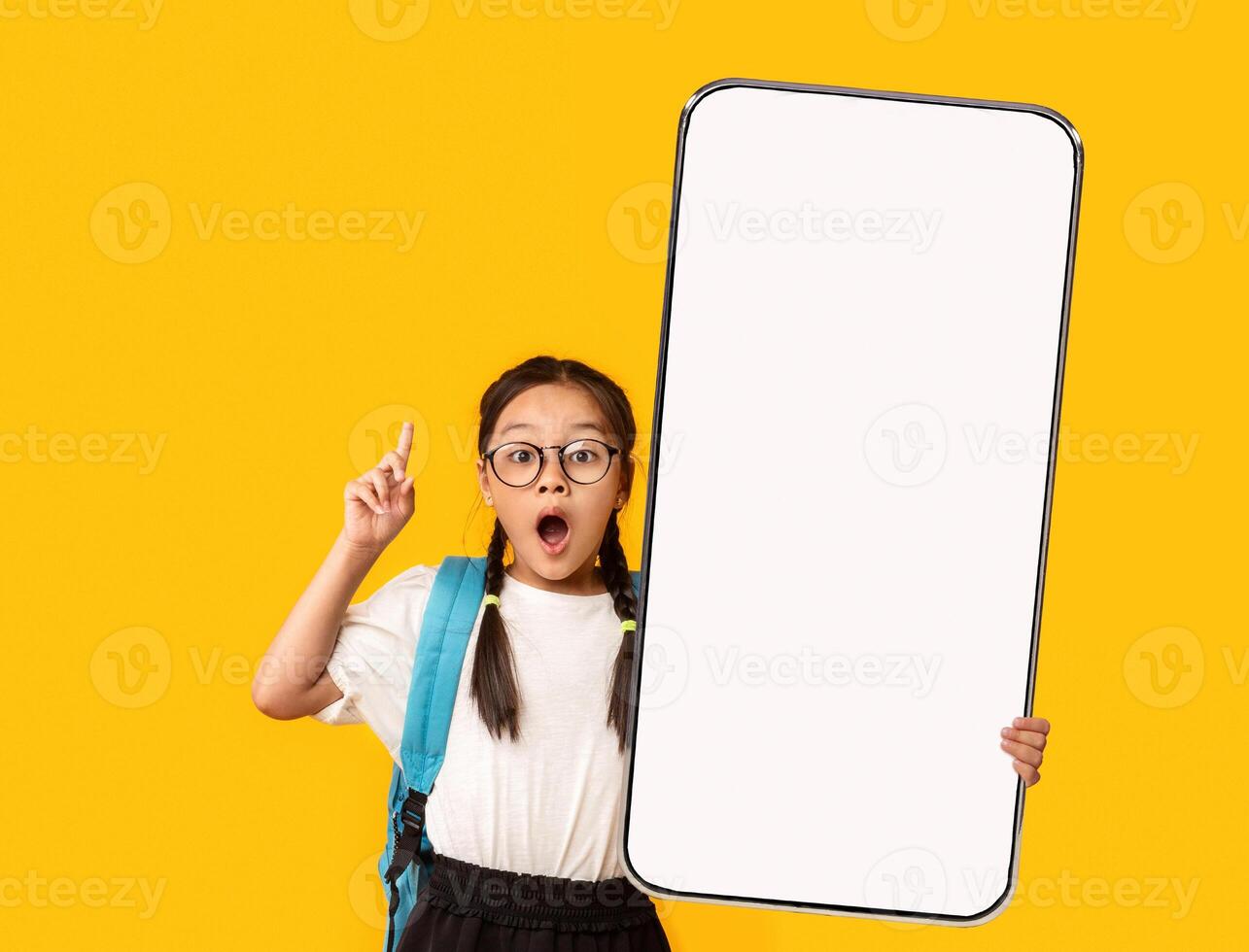 Wow, Great Offer. Excited Little Asian School Girl In Eyeglasses Holding Big Cell In Hand Pointing Finger Up Presenting Phone Empty Screen On Yellow Orange Studio Background. Smartphone Display Mockup photo
