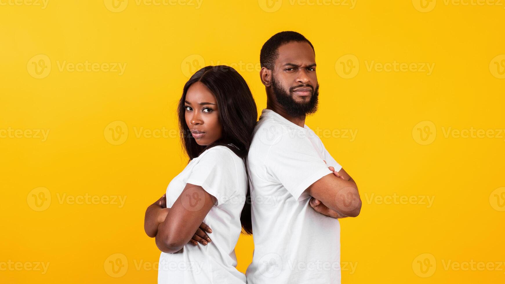 He vs She. Portrait of serious African American young couple standing back to back with folded arms, boyfriend and girlfriend looking at camera ith crossed hands isolated over yellow studio background photo