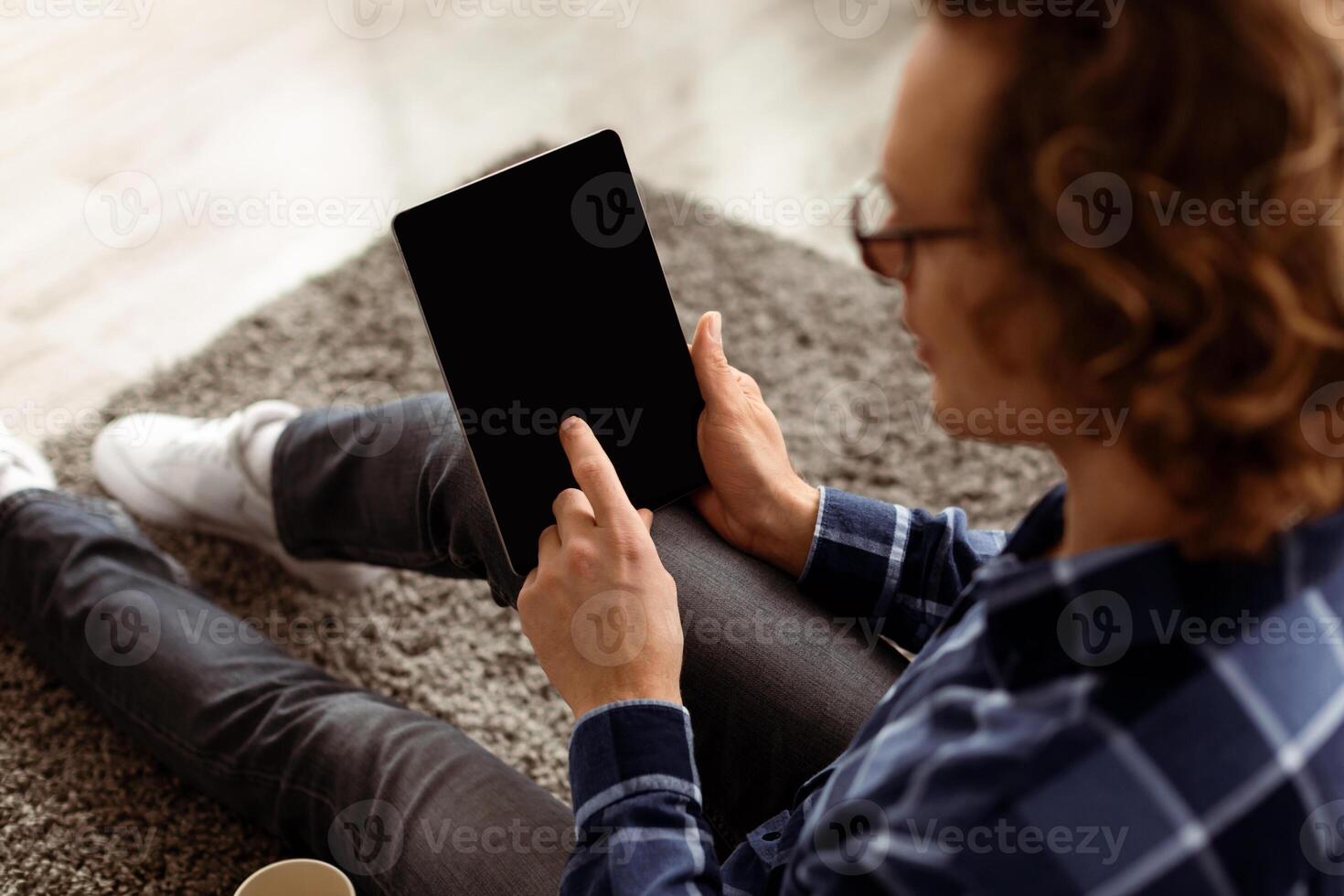 Unrecognizable Man Using Digital Tablet With Blank Screen For Application Or Website Advertisement. Back View Of Guy Browsing Internet On Computer Sitting On Floor At Home. Mockup, High Angle photo