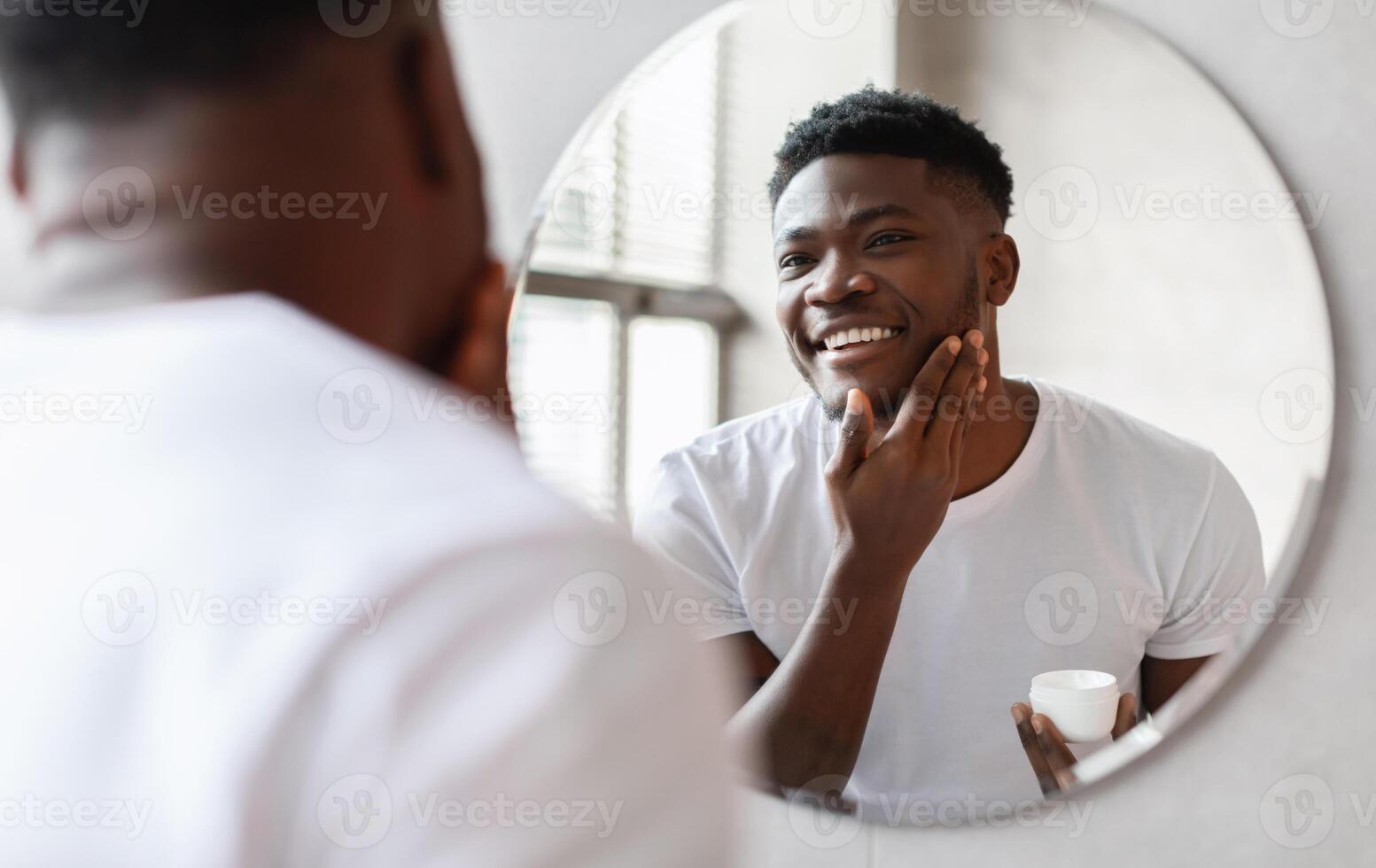 Smiling African American Man Touching Face Applying Moisturizer On Face Holding Moisturizing Cream Jar Looking At His Reflection In Round Mirror Standing In Bathroom At Home. Facial Skincare photo