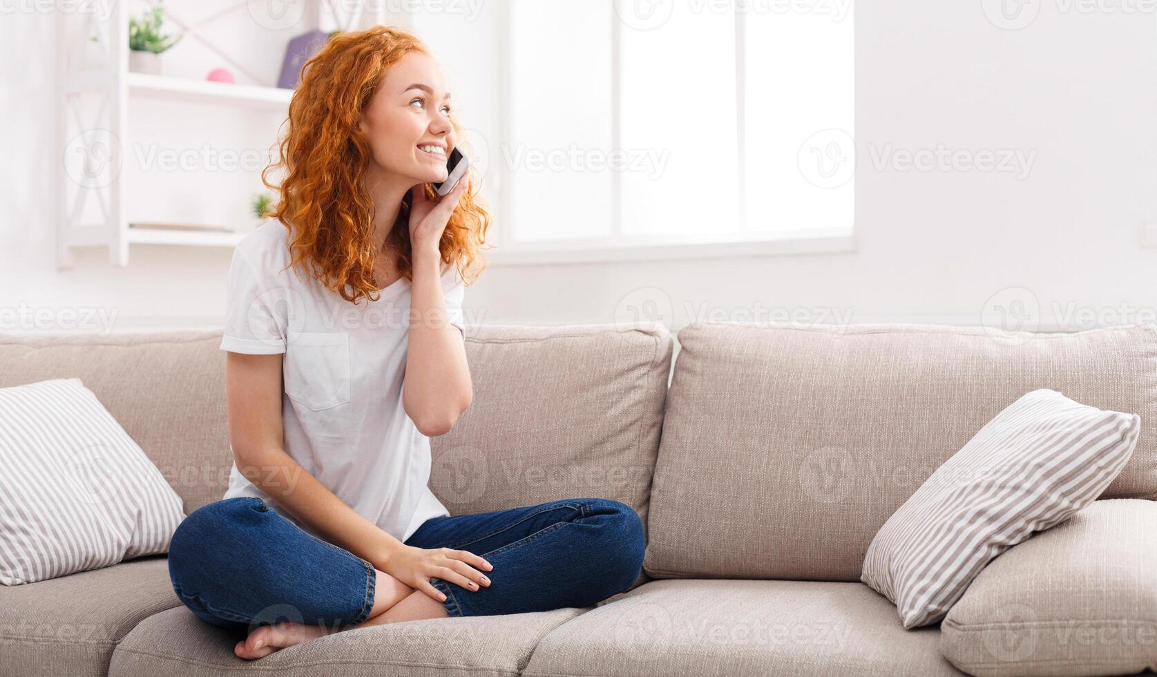 Pleasant talk with friend. Attractive smiling young redhead woman talking on cell phone while sitting on the beige sofa at home. Technology, communication and coziness concept photo