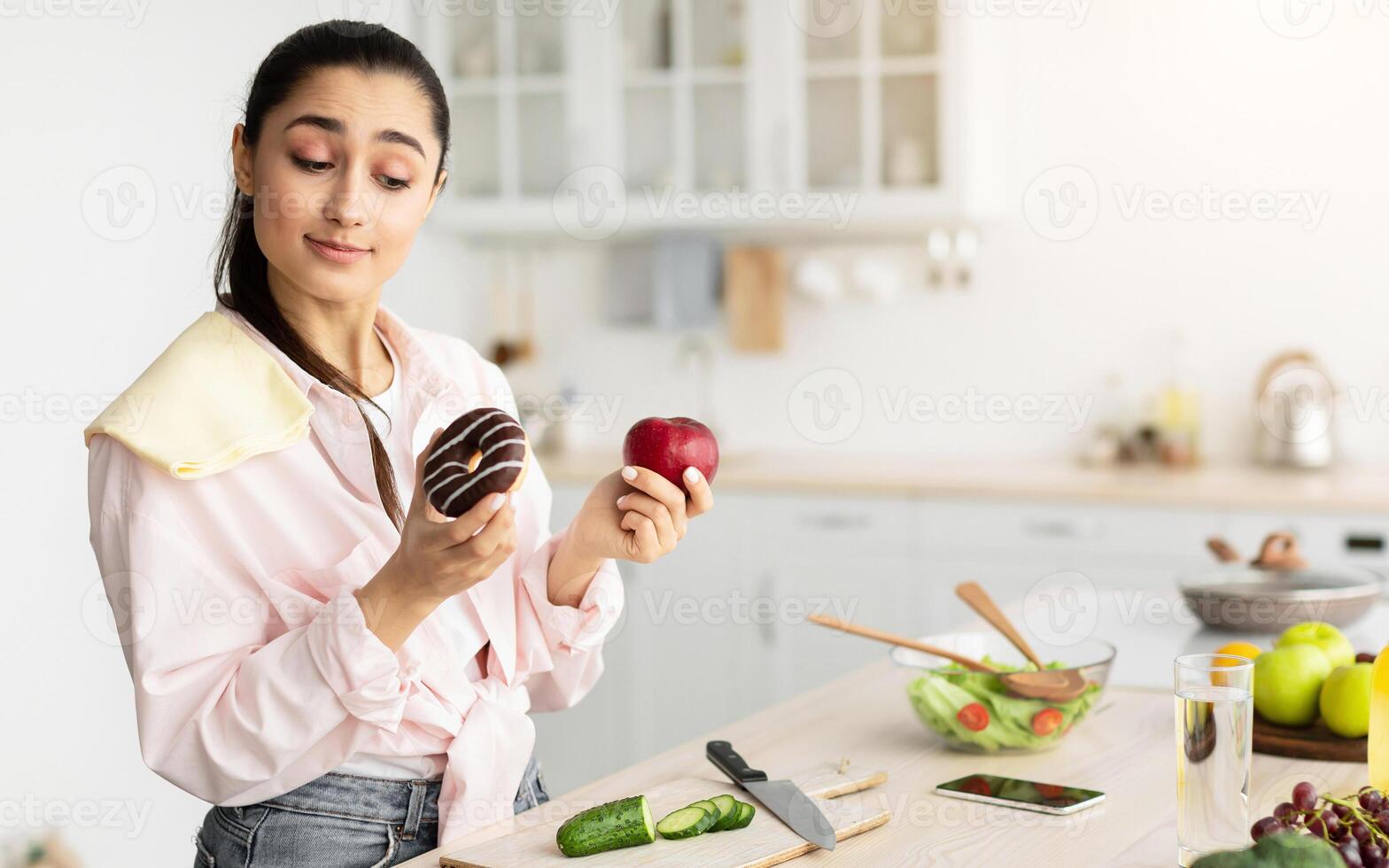 Healthy vs Unhealthy Food. Decisive doubtful woman holding red apple and donut in both hands, making choice of her meal, standing in kitchen, free copy space. Uncertain lady hesitating about her diet photo