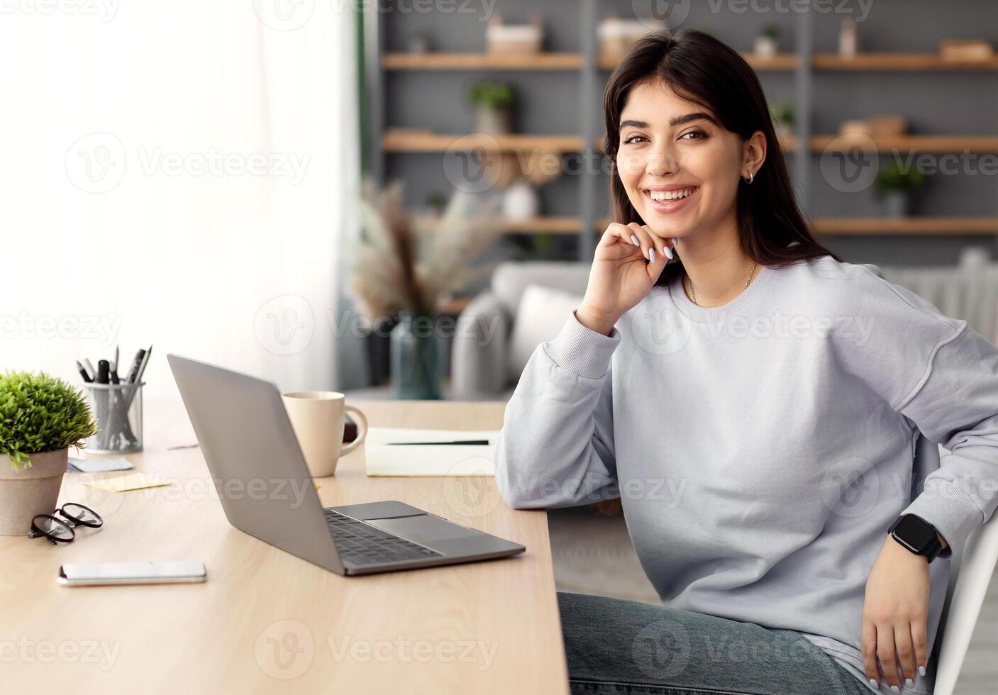 Professional Worker. Portrait of smiling young woman sitting at desk at home office, posing and looking at camera, working on laptop computer in workplace or living room, free copy space photo