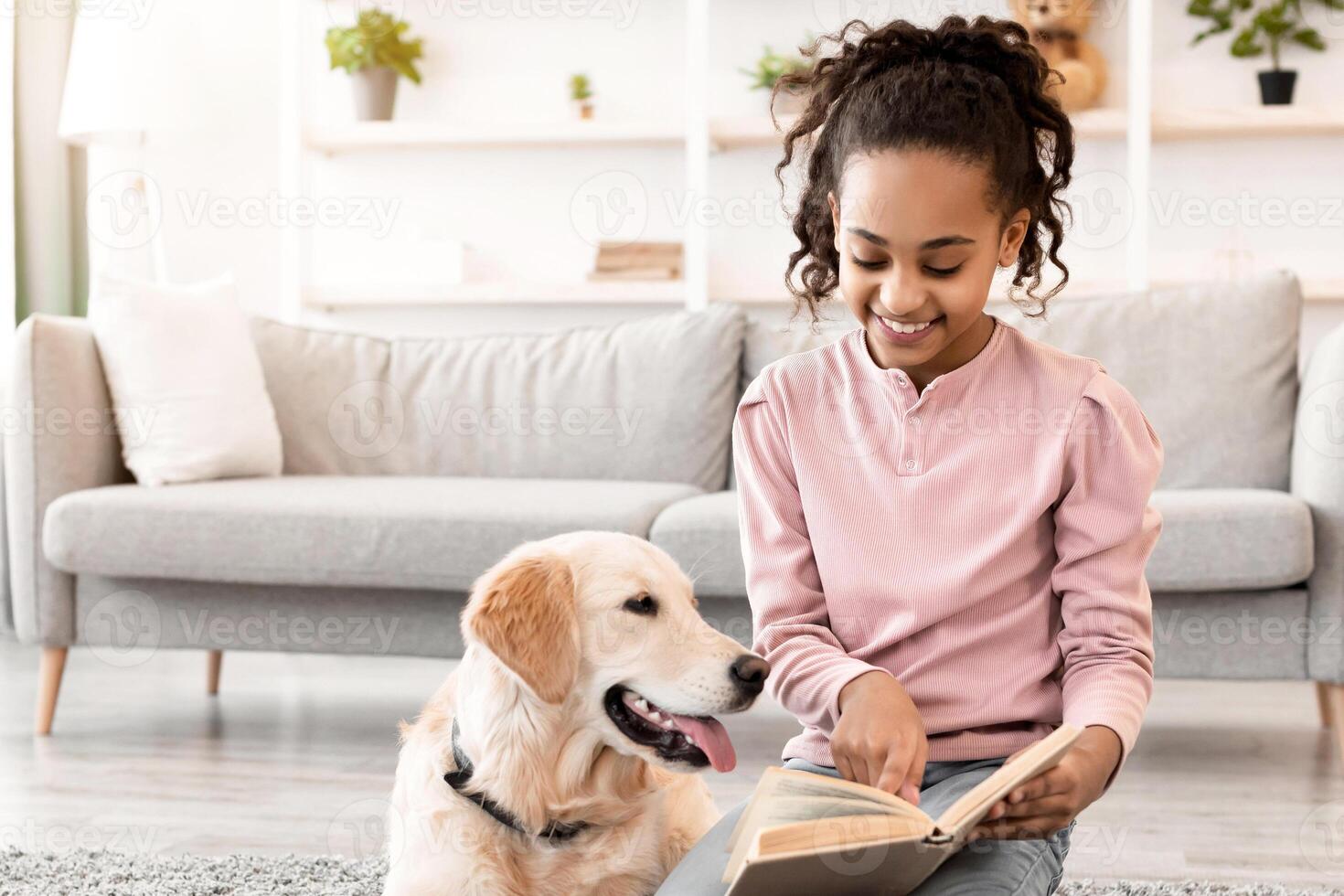 Leisure Time Concept. Portrait of cheerful black girl reading paper book and showing page to dog, sitting on the floor carpet with golden retiever in living room or bedroom at home, having fun photo
