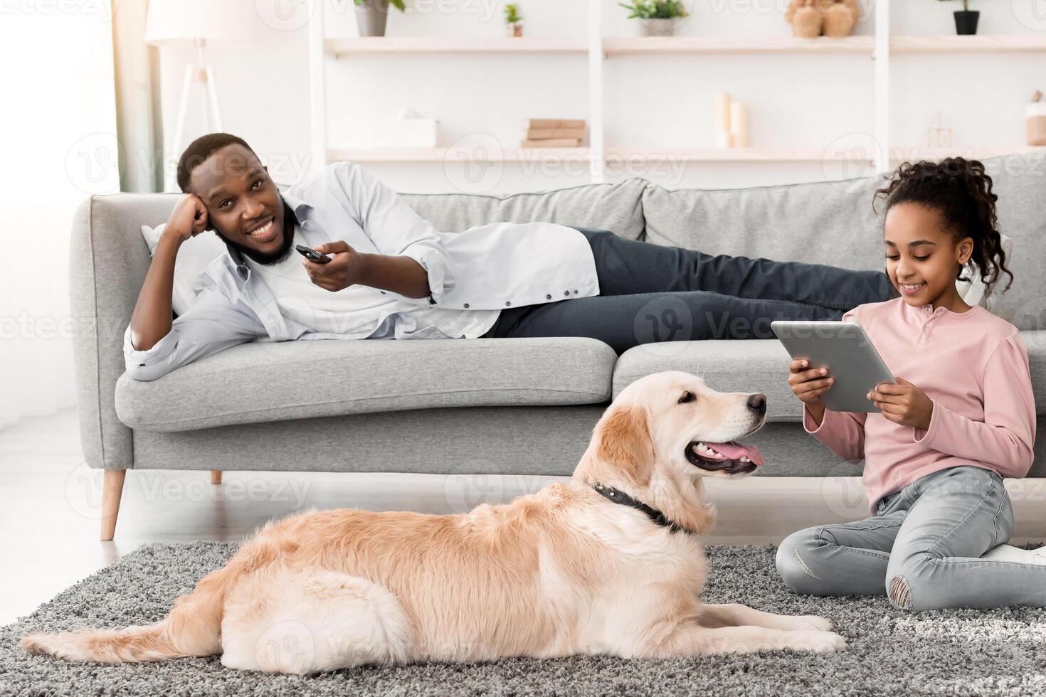 Weekend Concept. Black man lying on couch, watching television, holding remote control, happy puppy lying on carpet, smiling girl sitting on floor using tablet spending time together. Family Life photo