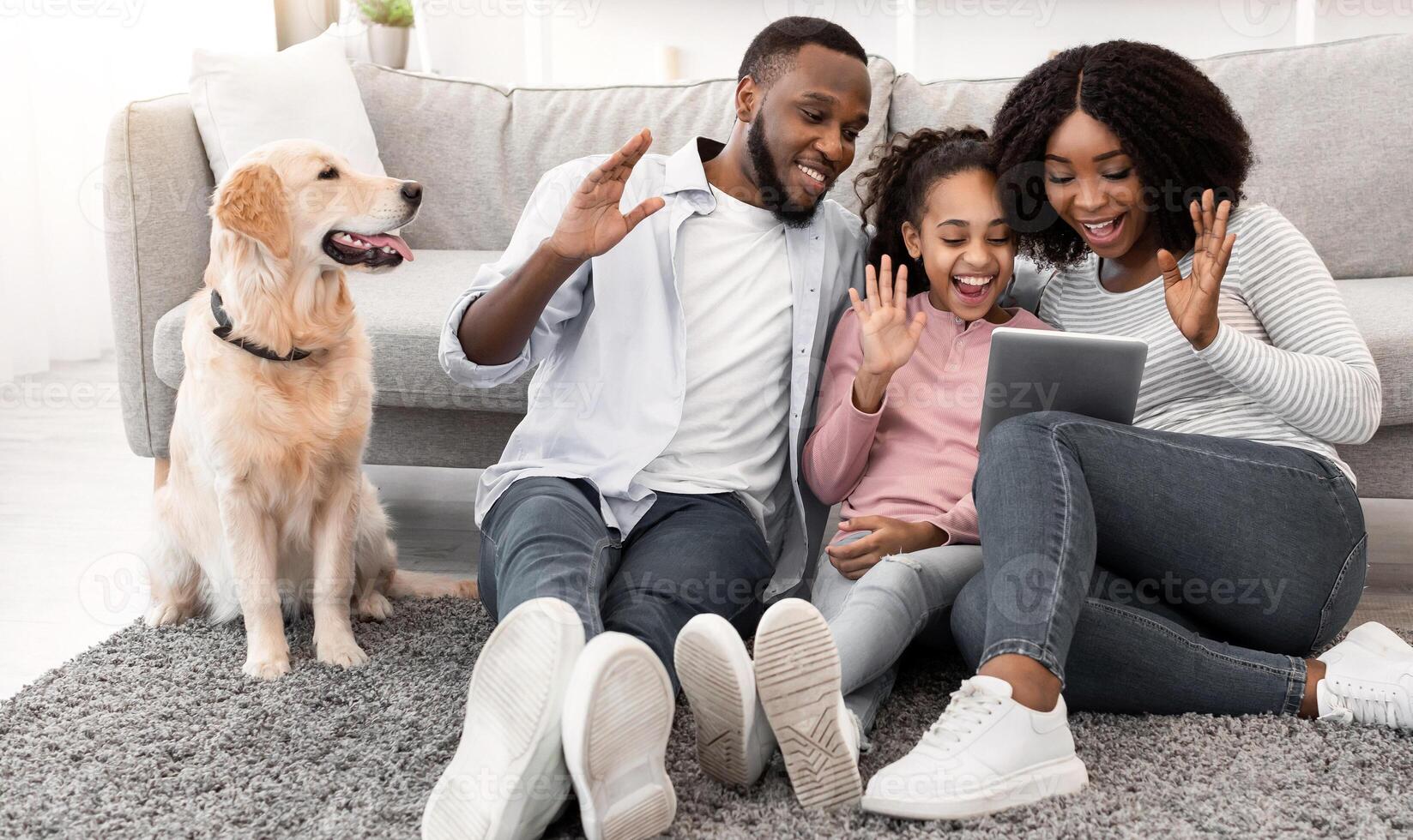 Online Communication. Smiling african american man, woman and girl making digital chat with friends or family using tablet, waving to webcam, sitting with dog on the floor carpet in living room photo