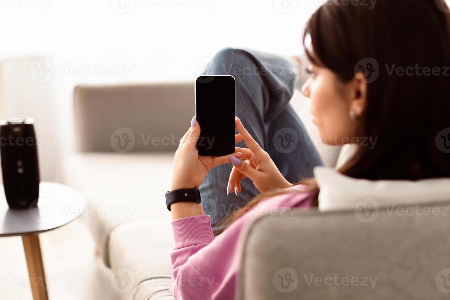Over The Shoulder View Of Unrecognizable Young Woman Holding And Using Cell Phone With Empty Mockup Screen, Sitting On Couch In Living Room At Home. Selective Focus On Gadget, Template photo