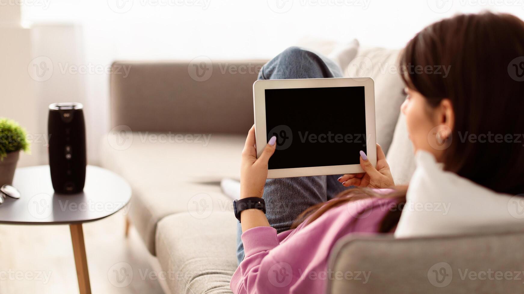 Over The Shoulder View Of Unrecognizable Young Woman Holding And Using Digital Tablet With Empty Mockup Screen, Sitting On Couch In Living Room At Home. Selective Focus On Gadget, Template photo