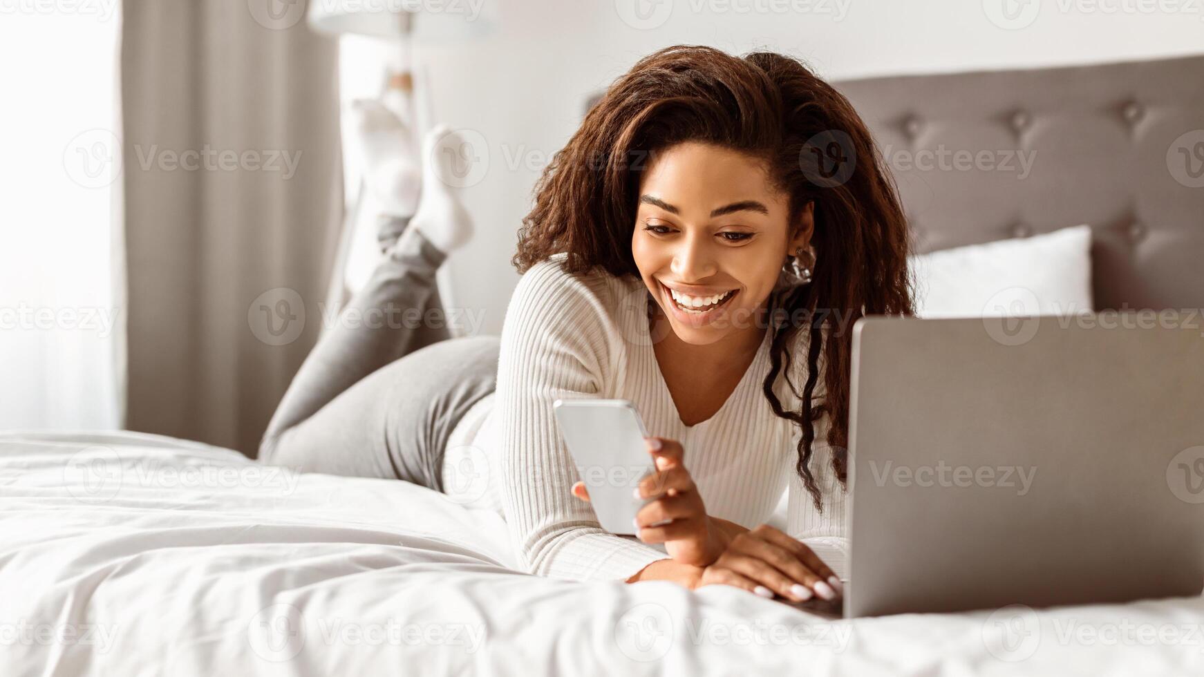 Portrait of excited African American woman lying in bed, reading text message with good news, holding and using mobile phone, working on laptop computer, black lady feeling happy, free copy space photo