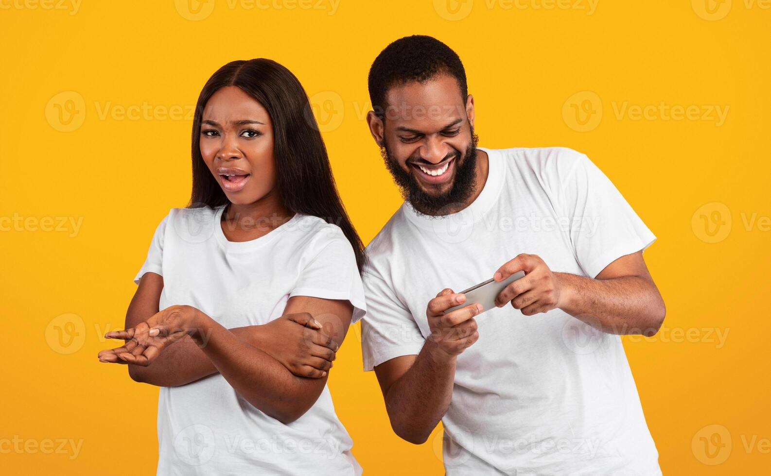 Phubbing. Excited black man stuck in smartphone, playing mobile games while angry young woman feeling unhappy about neglect and lack of communication. Jealous lady and gadget addicted man at studio photo