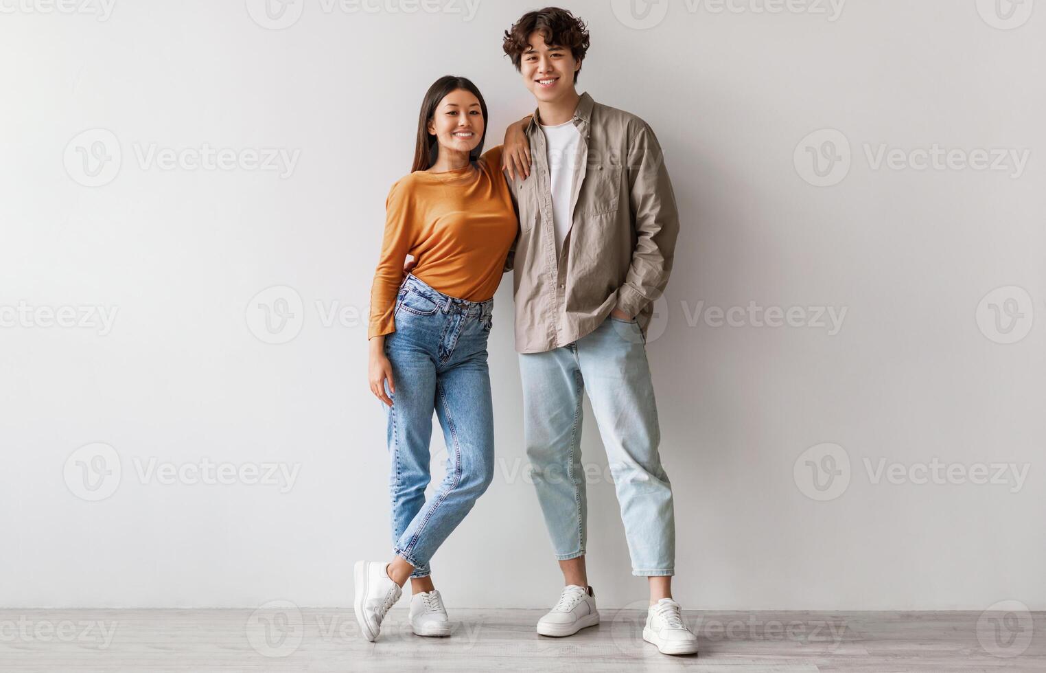 Full length portrait of happy young Asian couple in casual clothes hugging and looking at camera against white studio wall. Cheerful millennial spouses posing and smiling together photo