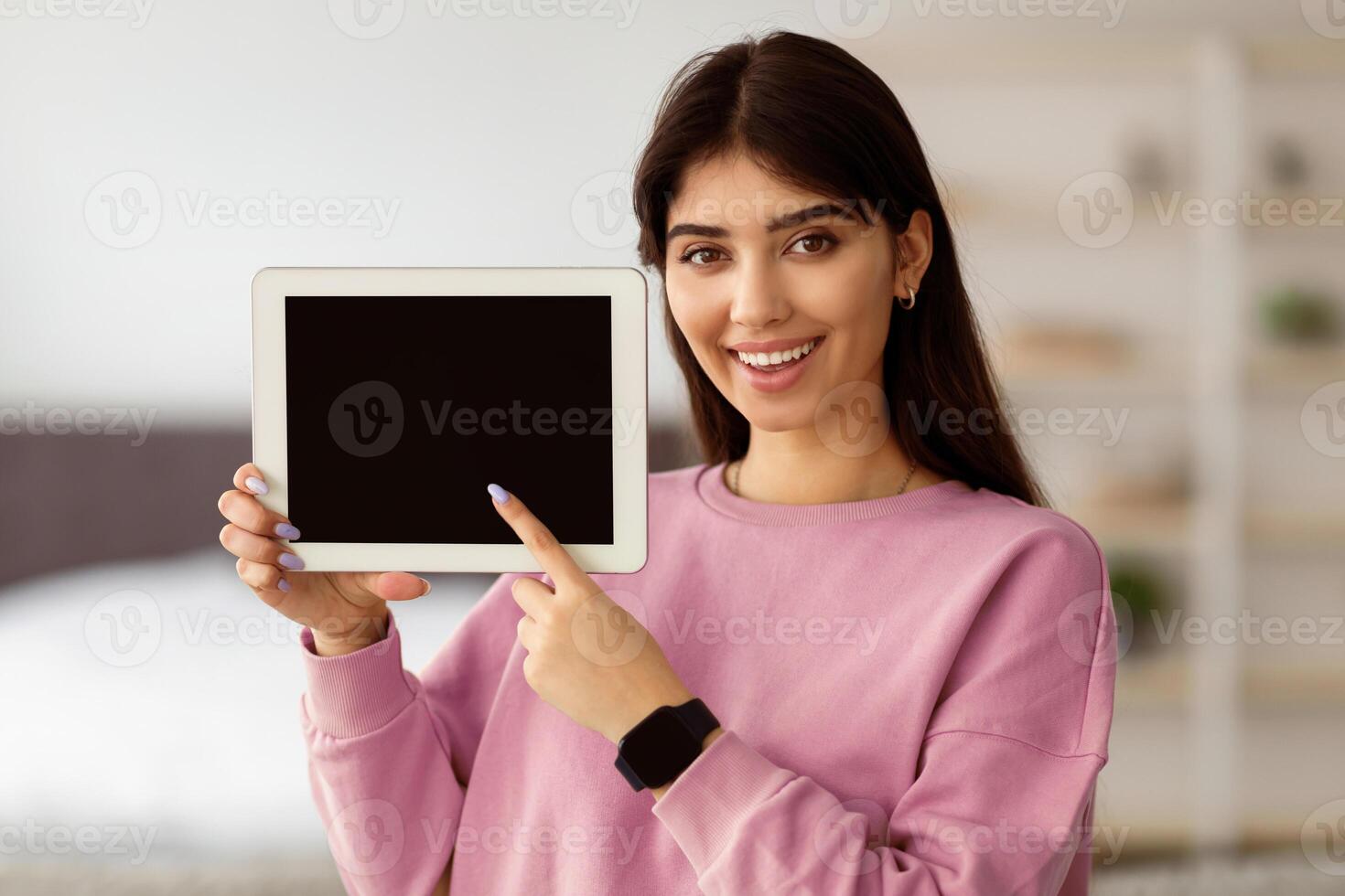 Great App. Portrait of excited young woman holding and showing digital tablet with black empty screen in hand, presenting and pointing at gadget screen with blank space for mock up, selective focus photo