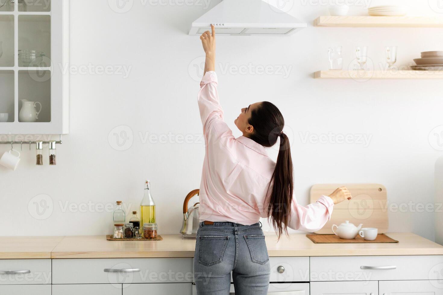 Household Concept. Back rear view of casual lady choosing and selecting mode on range exhaust hood, pushing button on mechanical fan above the stove. Female standing cooking in modern kitchen at home photo