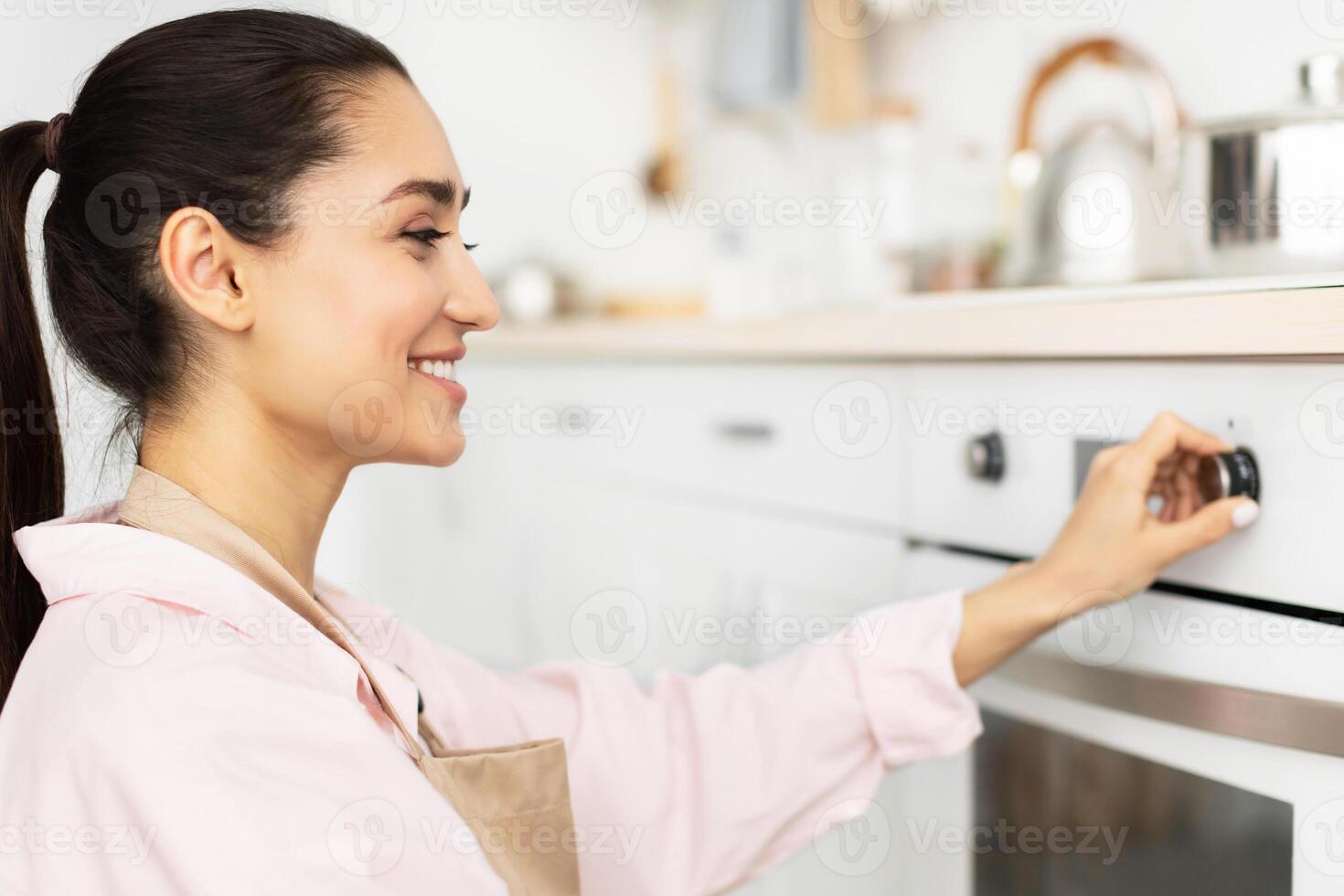 Closeup side view profile portrait of young smiling housewife in apron using stove in modern kitchen at home, holding and turning knob, controlling temperature of the burner, preparing meal photo