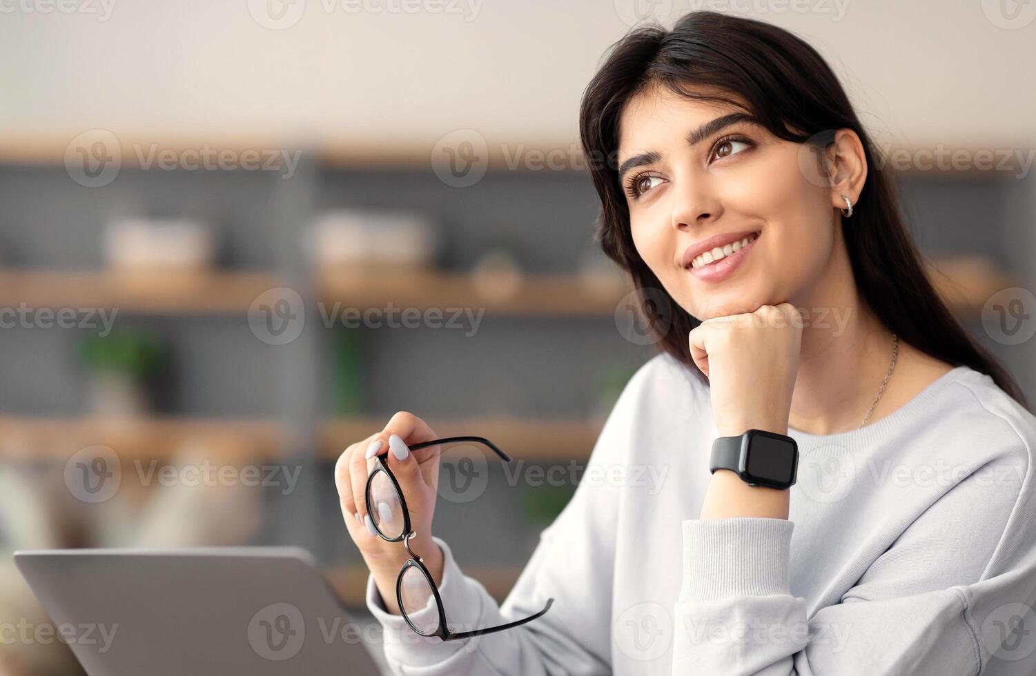 Photo of thinking young woman sitting and desk, holding eyeglasses and dreaming, using laptop computer. Female manager or student looking aside, selective focus, blurred background, free space