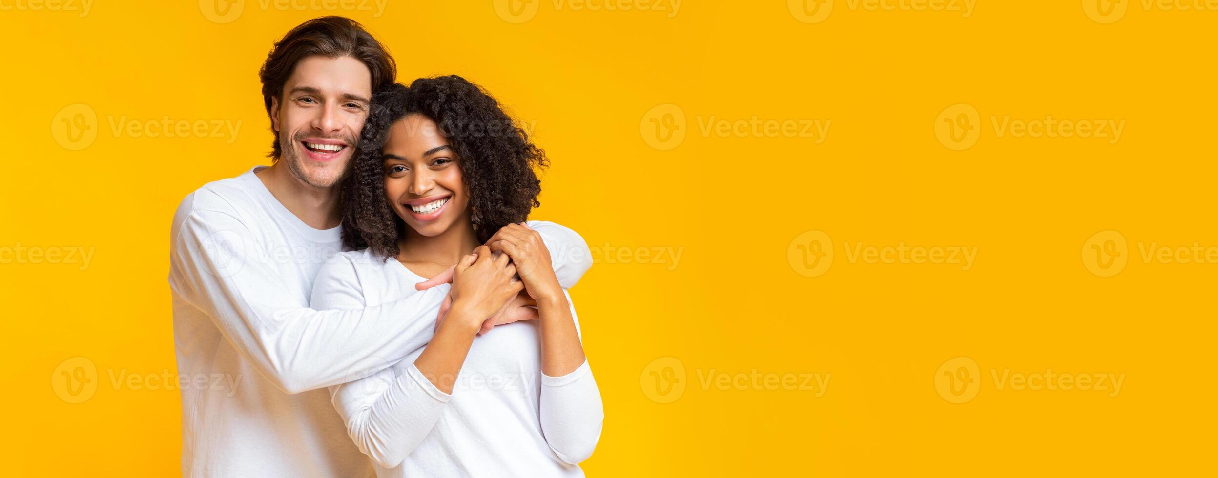 Happy interracial couple portrait. Joyful mixed-race sweethearts embracing and posing to camera, standing together on yellow background, panorama photo