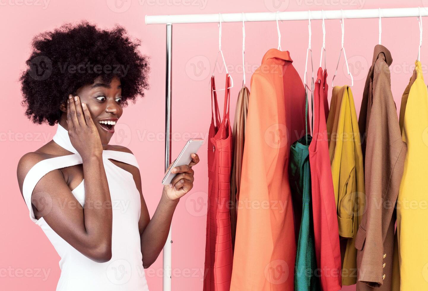 Great Shopping Offer. Excited African American Woman Using Phone With Shopper App Reading News About Sales And Discounts In Smartphone Standing Near Clothing Rack On Pink Background In Studio photo