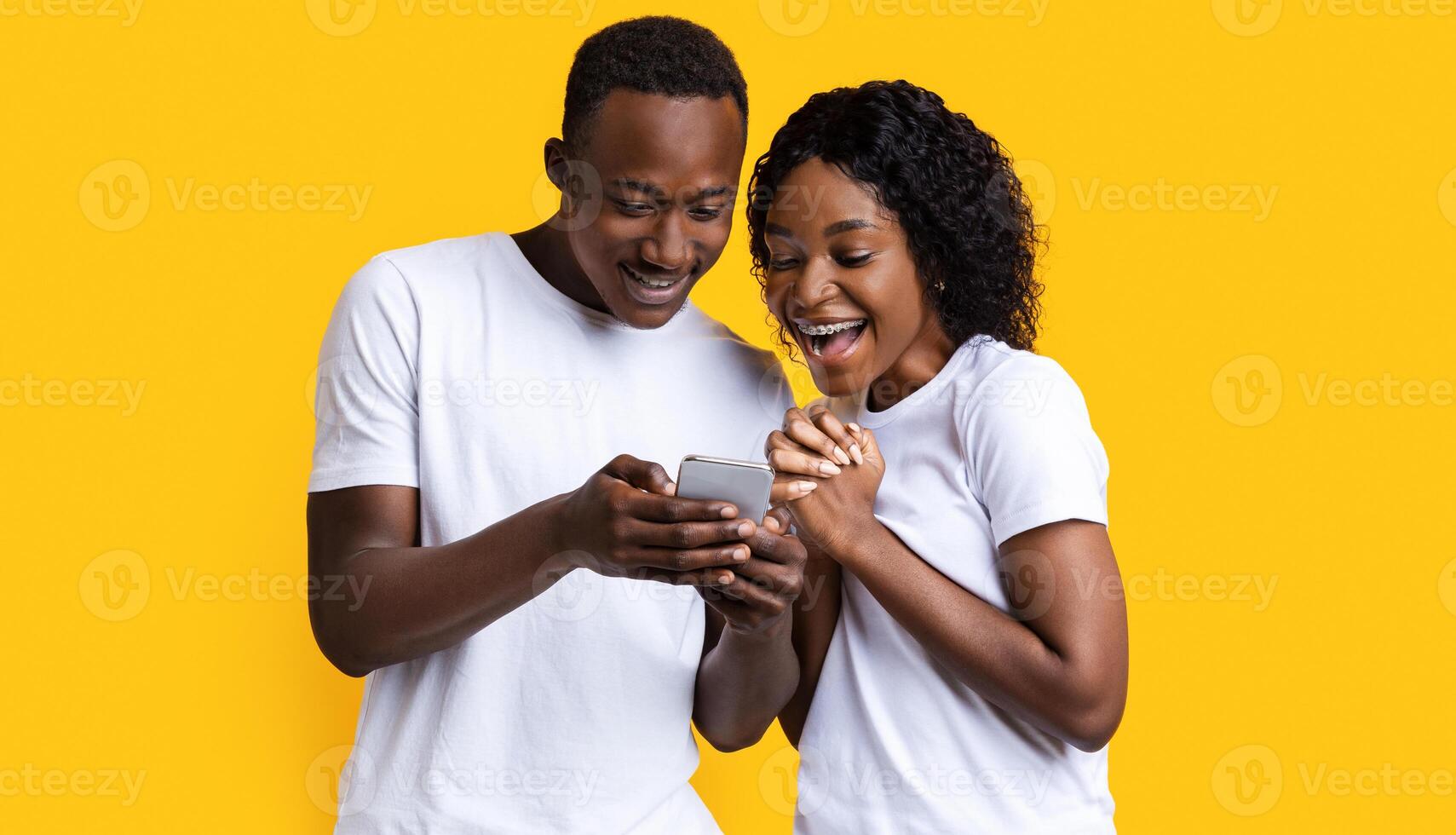 Excited black couple looking at mobile phone screen, using new cool application, yellow studio background. Joyful african american man and woman using smartphone together, entertainment concept photo