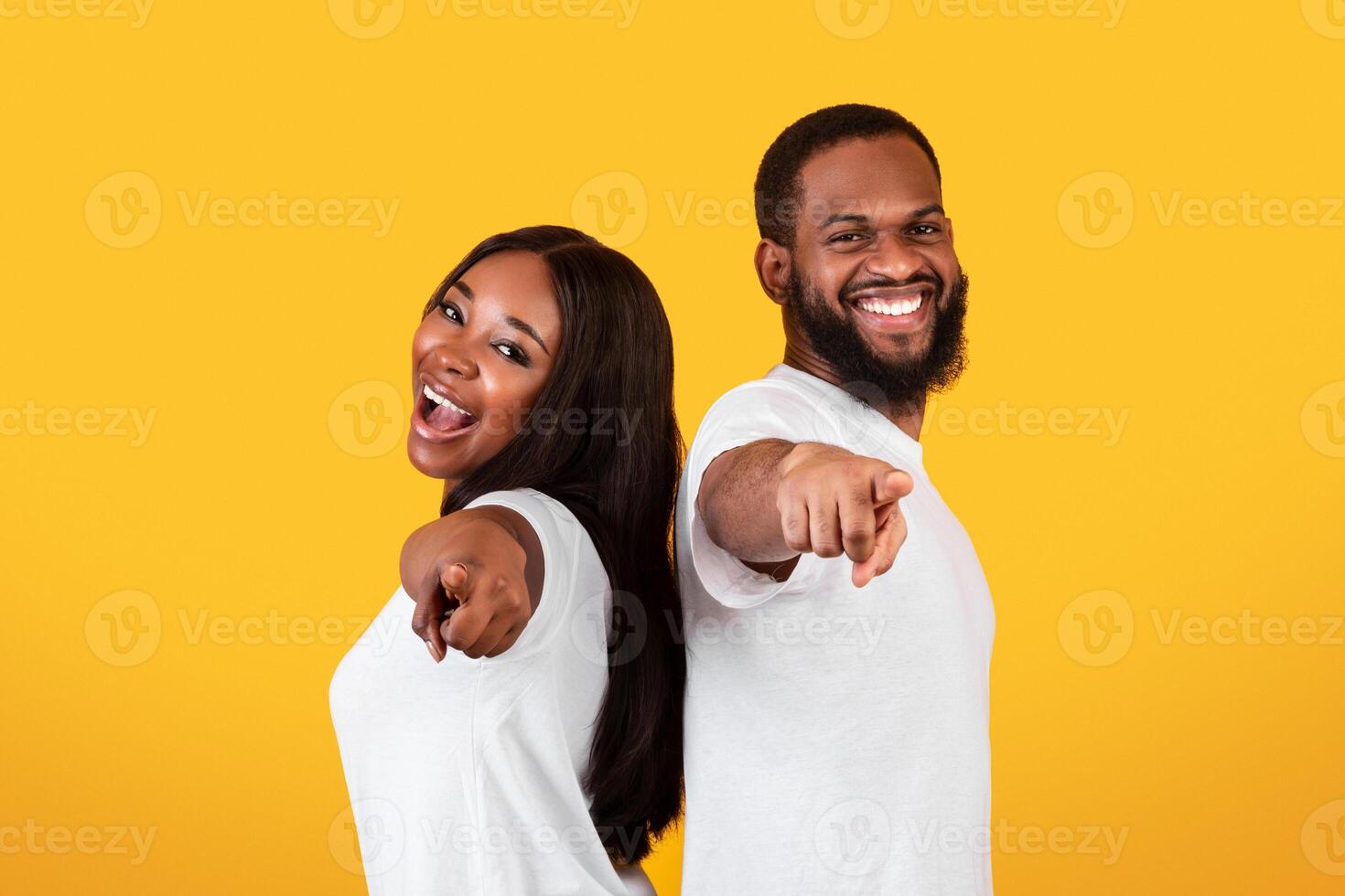 Your Turn. Portrait Of Excited African American Young Couple Pointing Fingers At Camera, Happy Man And Woman Choosing You, Standing Back To Back Isolated Over Yellow Studio Background, Copy Space photo